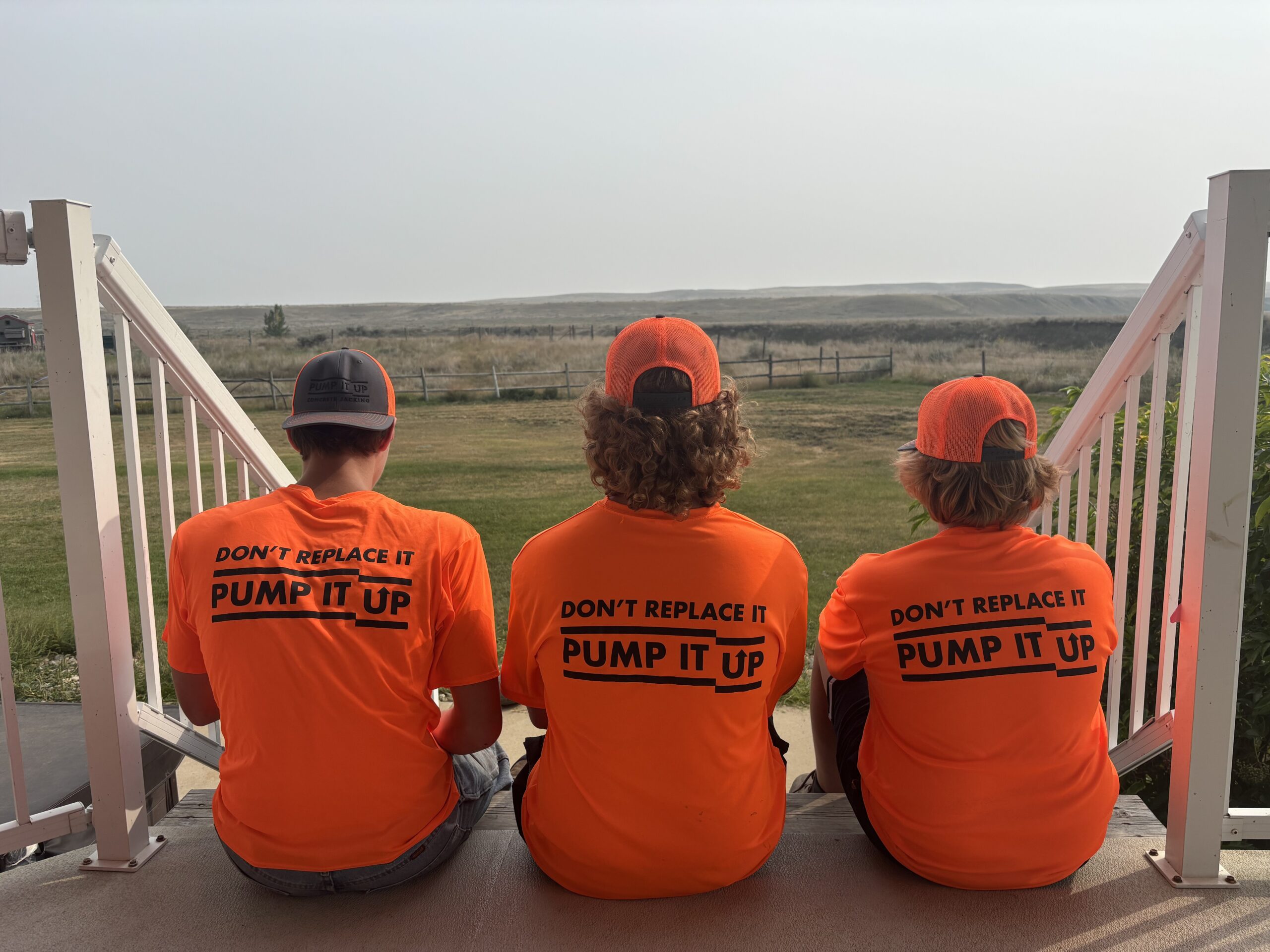 Three Pump It Up Concrete Jacking crew members in bright orange shirts sitting on steps overlooking Medicine Hat